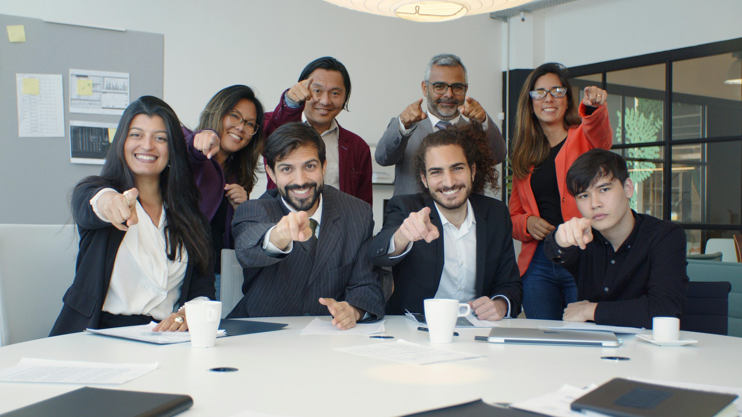 A diverse group of colleagues smiling and pointing in a modern office setting, promoting teamwork.
